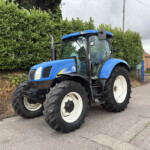 Blue New Holland tractor parked on a street beside a tall hedge under a cloudy sky.