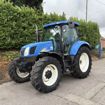 Blue New Holland tractor parked on a street beside a tall hedge under a cloudy sky.