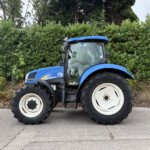Blue New Holland tractor parked on a paved surface with a tall green hedge behind it.