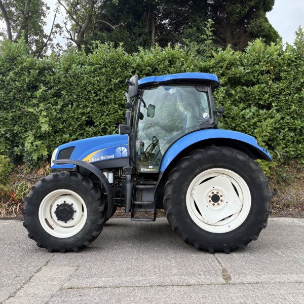 Blue New Holland tractor parked on a paved surface with a tall green hedge behind it.