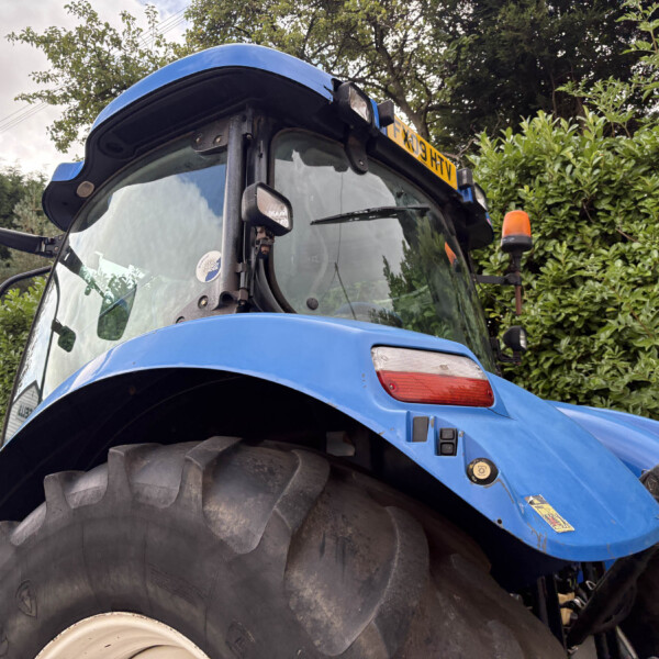 Close-up of a blue tractor front with oversized treaded tire and orange beacon, greenery in background, angled from the left side
