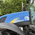 Close-up of a blue New Holland tractor cab with exhaust stack, front fender, and a green hedge in the background.