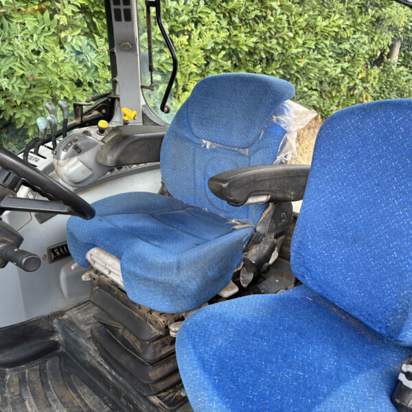 Inside a construction vehicle cabin with two blue upholstered seats, one wrapped in plastic, and dusty, worn controls and floor.