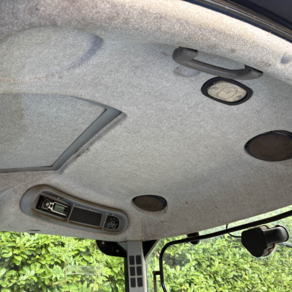 Interior of a vehicle cabin ceiling with a grab handle, overhead light, and control panel; greenery visible through the window outside.