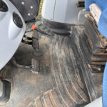 Dusty vehicle cab floor with three pedals (clutch, brake, accelerator) and a dirty rubber mat; blue fabric seat visible at right edge.