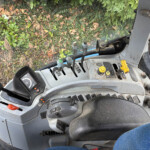 Inside a tractor cab showing gear levers, control sticks, and a dusty dashboard with green plants outside the window.