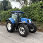 Blue New Holland tractor parked on a driveway beside a tall green hedge, with a small building and orange beacon on top in the background.