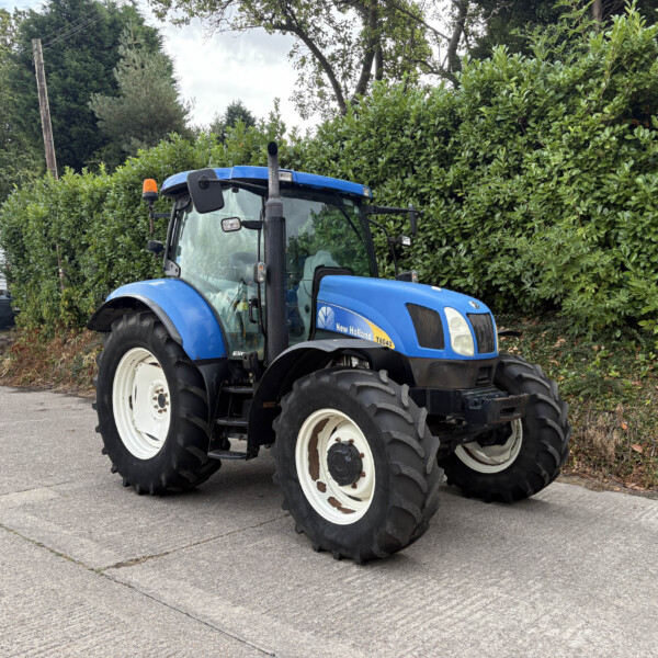 Blue New Holland tractor parked on a driveway beside a tall green hedge, with a small building and orange beacon on top in the background.