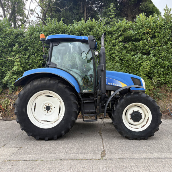 Blue New Holland tractor parked on a paved surface with a tall green hedge in the background, front cab visible.