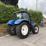 Blue tractor parked on a street beside a tall green hedge with brick houses in the background, on a cloudy day.