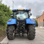 Rear view of a large blue tractor with oversized tires, parked on a road beside a tall hedge and brick houses under a cloudy sky.