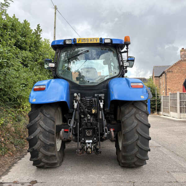 Rear view of a large blue tractor with oversized tires, parked on a road beside a tall hedge and brick houses under a cloudy sky.