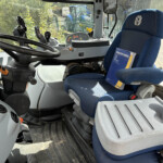 Interior of a tractor cab with a large steering wheel, pedals, and a blue driver seat; an operator's manual sits on the seat edge.