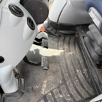 Interior of a vehicle cab showing three floor pedals and a bright orange ribbed knob near them; the floor is dusty and rubberized with grooves.