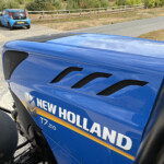 Close-up of a blue New Holland tractor hood with vents, parked in a lot beside a white car and a wooden fence in the background.