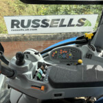 Interior view of a tractor cab with numerous control levers and colorful buttons on a gray dashboard, a window sign reading 'RUSSELLS' outside.