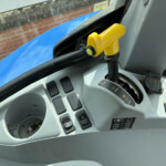 Yellow joystick control in a vehicle cab, mounted on a black lever with a gray dashboard and blue exterior visible through the window outside.
