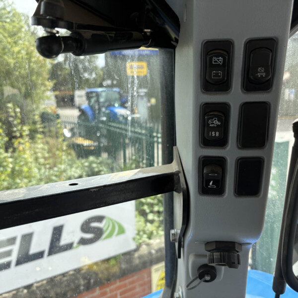 Interior of a vehicle cab with a vertical right-side control panel and multiple switches; outside view through a dirty windshield shows trees and a street, plus an emergency exit sticker on the left window.