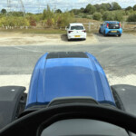 View from inside a blue car looking at two parked cars (white and blue) in a gravel lot with trees and a fence in the background, dashboard visible.