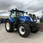 Blue New Holland tractor with large white wheels parked in a yard, with other tractors and a brick wall behind in overcast light.