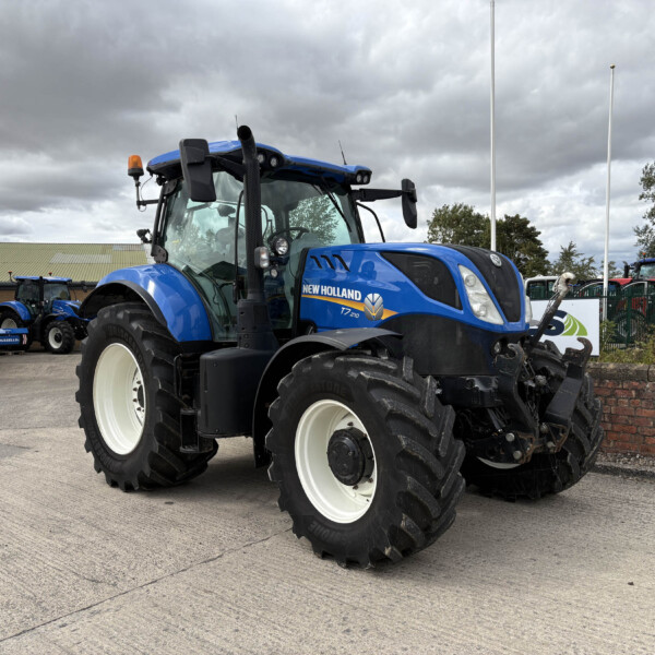 Blue New Holland tractor with large white wheels parked in a yard, with other tractors and a brick wall behind in overcast light.