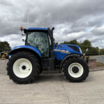 Blue New Holland tractor in a parking lot with other farm machinery, facing left under a cloudy sky, showing large white wheels and cab windows.