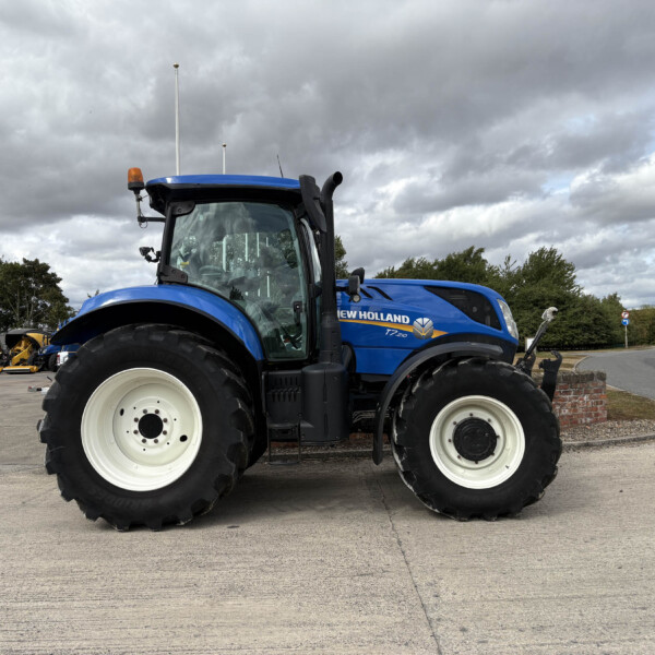Blue New Holland tractor in a parking lot with other farm machinery, facing left under a cloudy sky, showing large white wheels and cab windows.