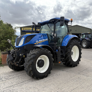Blue New Holland tractors parked outside a dealership under a cloudy sky, with a sign reading 'New Holland Agriculture' in the background.