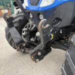 Close-up of a blue tractor front showing a rusted hitch, hydraulic arms, and large dirty tires in a yard setting.