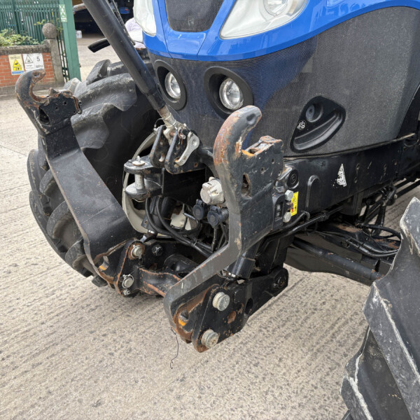 Close-up of a blue tractor front showing a rusted hitch, hydraulic arms, and large dirty tires in a yard setting.