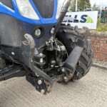 Close-up of a blue tractor's front hydraulic hitch and arms, with rusted metal parts and multiple hoses, on a concrete surface near a brick wall.