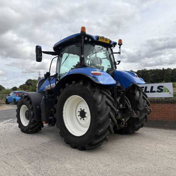 Blue New Holland tractor parked in a lot under a cloudy sky, large white-rimmed wheels visible.