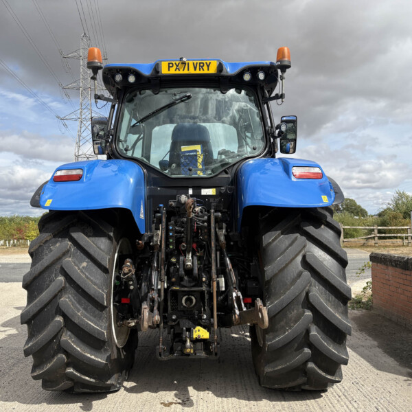 Front view of a blue tractor with oversized rear tires and orange warning lights, license plate PX71 VRY, under a cloudy sky at a rural site.