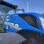 Close-up of a blue New Holland T7.210 tractor front, showing the grille, headlight, and badge on a sunny day