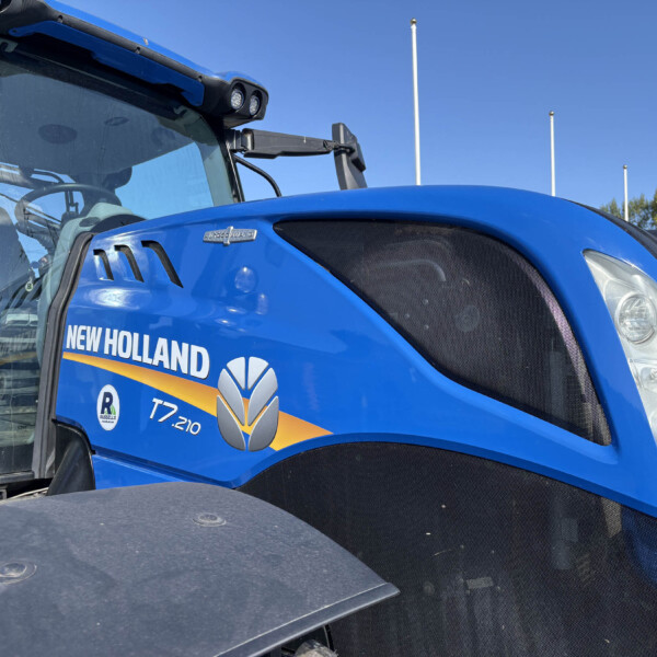 Close-up of a blue New Holland T7.210 tractor front, showing the grille, headlight, and badge on a sunny day