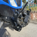 Close-up of a blue tractor front with hydraulic arms and large black tires, brick wall and fence in the background.