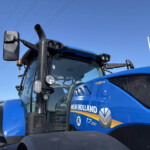 Close-up of a blue New Holland tractor cab with a tall perforated exhaust stack and side mirror against a clear blue sky.