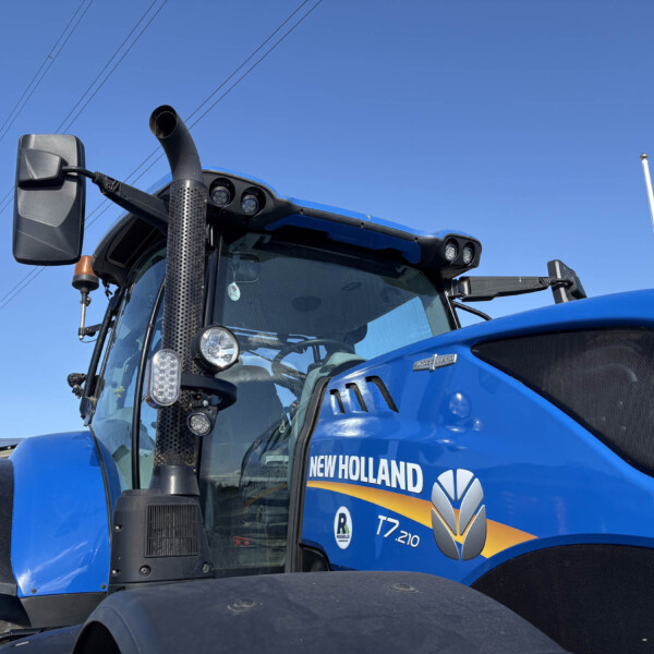 Close-up of a blue New Holland tractor cab with a tall perforated exhaust stack and side mirror against a clear blue sky.
