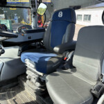 Inside the cab of a New Holland agricultural tractor: blue and gray seats, a multi-button control panel, steering wheel, and a view of farm buildings outside the windows.
