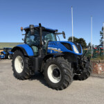 Blue New Holland tractor parked outdoors with large front tires and a clear blue sky, other farm equipment in the background