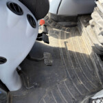 Close-up of a vehicle interior footwell showing two black textured pedals and a red knob, with a dirty rubber floor mat and grey dashboard panel nearby.