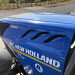 Close-up of a blue New Holland tractor hood with vents and a small Autocommand badge, rural fence and road in the background