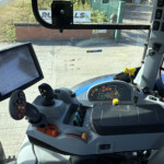 Inside a tractor cab showing a touchscreen display, joystick controls, and a full panel of buttons and levers nearby.