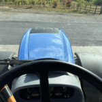 View from the driver's seat of a blue tractor, dashboard in foreground and road ahead with a fence and trees in the distance; windshield is dusty.