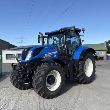 Blue New Holland tractor with large front tires parked in an outdoor yard near green fencing and industrial buildings under a clear blue sky.