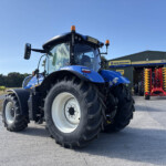 Close-up of a blue New Holland tractor at an agricultural equipment yard, with a green metal building and sign reading New Holland Agriculture in the background.