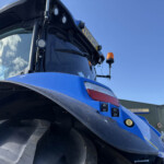 Close-up of a blue tractor front with a large tire, cab window, amber beacon and control buttons visible on the side side panel.