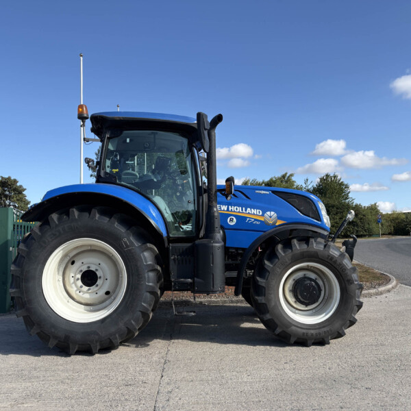 Front half of a blue New Holland tractor with large tires parked outdoors under a bright blue sky.