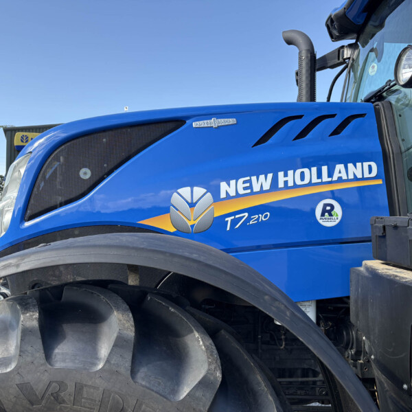 Blue New Holland tractor cab with oversized front tire and side branding visible (T7.210).