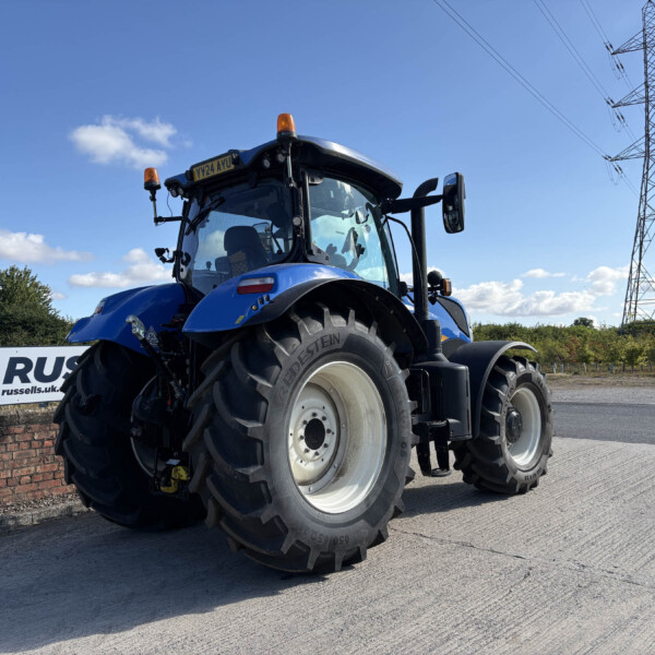 Front view of a blue tractor with large tires and orange beacon lights, beside a brick wall under a blue sky.